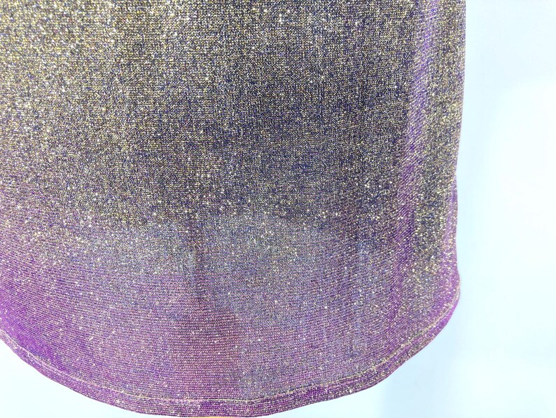 May include: Close-up of a shimmering fabric with a gradient effect. The top portion is a blend of gold and silver, transitioning to a deep purple at the bottom. The fabric has a textured, glittery appearance.