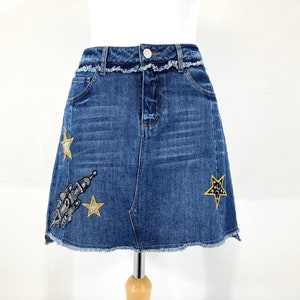 Pode incluir: Uma saia de ganga azul com bainha desfiada e bordados de estrelas douradas e foguetes.