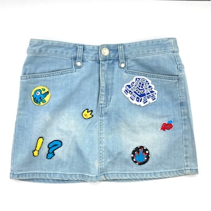 May include: A light blue denim mini skirt with colourful embroidered patches. The patches include a blue and yellow creature, a yellow crown, a blue and white building, a red heart, a blue and white swirl, and a blue and white exclamation mark.