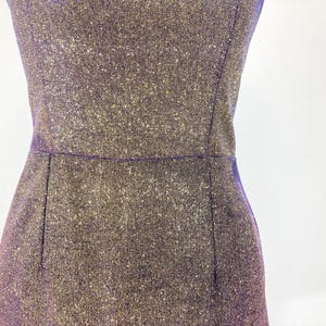 May include: Close-up of a shimmering, sleeveless dress in shades of purple and gold. The dress has a fitted silhouette with a defined waistline and a subtle sparkle. The fabric appears to be textured, catching the light.