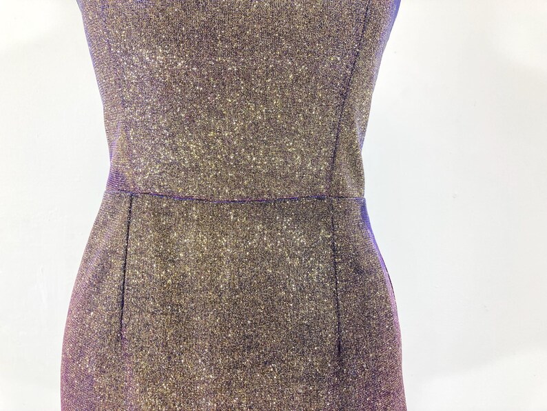 May include: Close-up of a shimmering, sleeveless dress in shades of purple and gold. The dress has a fitted silhouette with a defined waistline and a subtle sparkle. The fabric appears to be textured, catching the light.