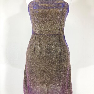 May include: A strapless, knee-length dress with a shimmering gold and purple fabric. The dress is fitted, with a straight neckline and a subtle A-line skirt. The fabric has a glittery texture, catching the light.