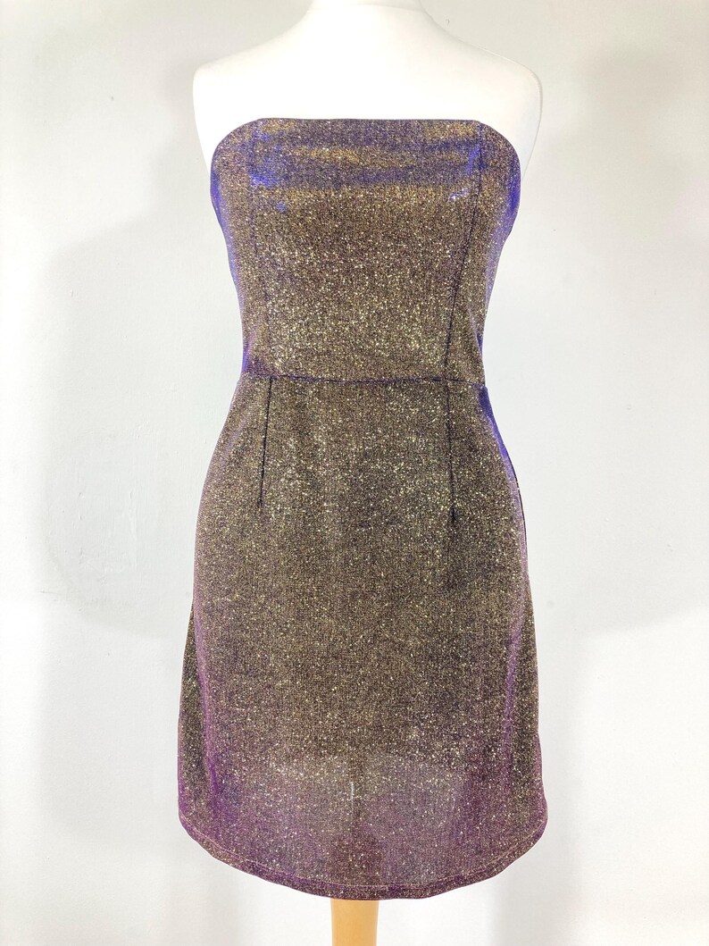 May include: A strapless, knee-length dress with a shimmering gold and purple fabric. The dress is fitted, with a straight neckline and a subtle A-line skirt. The fabric has a glittery texture, catching the light.