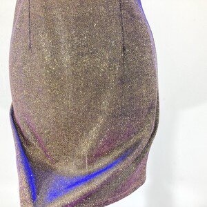 May include: Close-up of a shimmering, form-fitting dress. The fabric is a blend of gold, purple, and blue, creating a sparkling effect. The dress has a draped design on the side, adding a touch of elegance. The overall style suggests a party or evening wear.