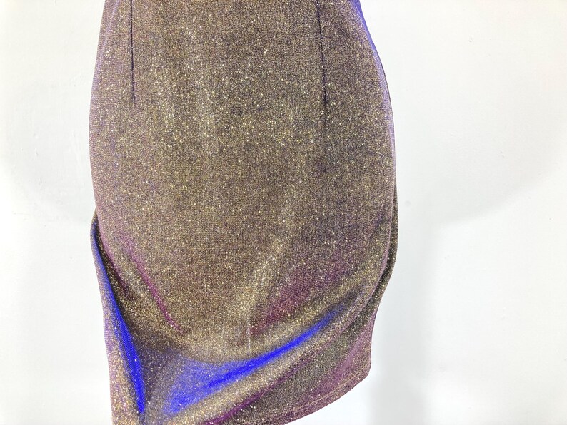 May include: Close-up of a shimmering, form-fitting dress. The fabric is a blend of gold, purple, and blue, creating a sparkling effect. The dress has a draped design on the side, adding a touch of elegance. The overall style suggests a party or evening wear.