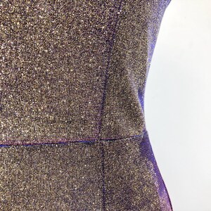 May include: Close-up of a shimmering, textured dress in shades of gold and purple. The fabric has a sparkly, glittery appearance. The dress has a fitted silhouette with a defined waistline and a simple, elegant design.