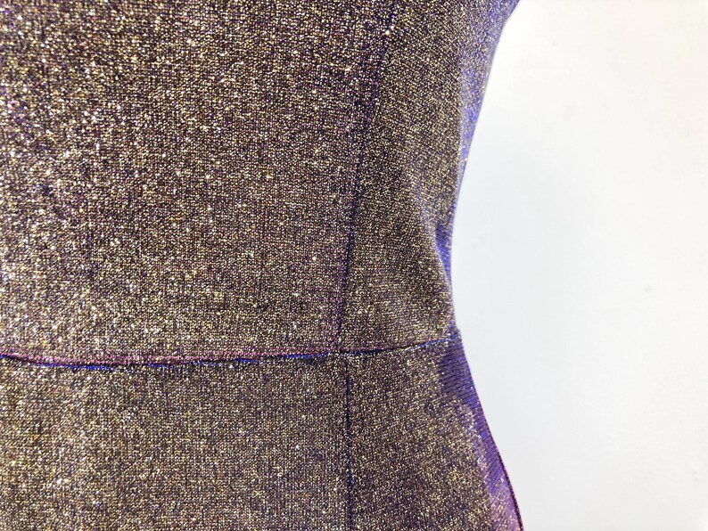 May include: Close-up of a shimmering, textured dress in shades of gold and purple. The fabric has a sparkly, glittery appearance. The dress has a fitted silhouette with a defined waistline and a simple, elegant design.