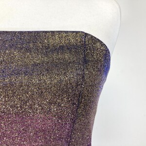May include: A strapless garment with a shimmering, textured fabric. The color gradient transitions from a deep purple at the bottom to gold and blue at the top. The fabric has a subtle sparkle, catching the light.