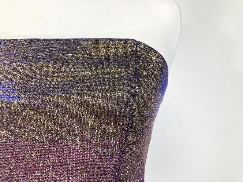 May include: A strapless garment with a shimmering, textured fabric. The color gradient transitions from a deep purple at the bottom to gold and blue at the top. The fabric has a subtle sparkle, catching the light.