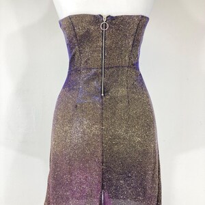 May include: A strapless, shimmering mini-dress with a gradient effect, transitioning from gold to purple. The dress features a fitted bodice with a zipper closure down the back. The fabric has a glittery texture.