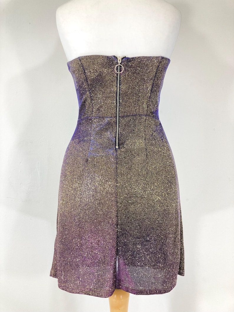 May include: A strapless, shimmering mini-dress with a gradient effect, transitioning from gold to purple. The dress features a fitted bodice with a zipper closure down the back. The fabric has a glittery texture.