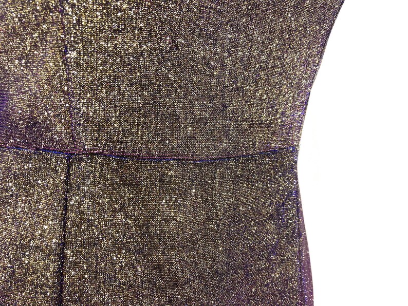 May include: Close-up of a shimmering, textured fabric in gold and black. The material appears to be a dress or garment, with a visible seam at the waist. The fabric has a sparkly, glittery appearance.