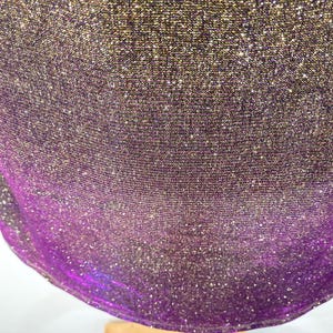May include: Close-up of a shimmering fabric with a gradient effect. The material transitions from a dark gold at the top to a vibrant purple at the bottom. The fabric has a textured appearance and is slightly draped.