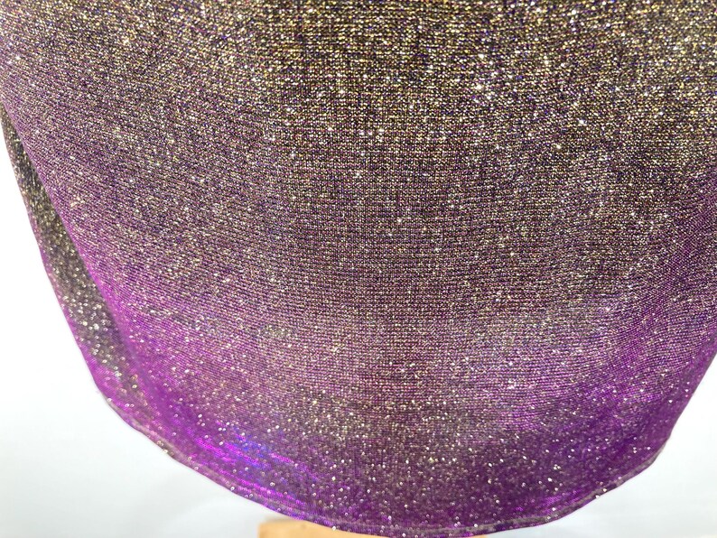 May include: Close-up of a shimmering fabric with a gradient effect. The material transitions from a dark gold at the top to a vibrant purple at the bottom. The fabric has a textured appearance and is slightly draped.