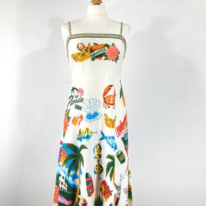 Pode incluir: Vestido branco de verão com estampa tropical com bananas, flores e o texto "Paradise". O vestido tem alças finas e um design colorido com palmeiras, pranchas de surf e o texto "The Paradise Inn."