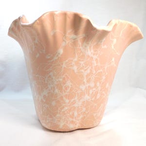 May include: A peach-colored ceramic vase with a ruffled rim and a white splatter design. The vase has a flared shape and a smooth, glossy finish. The decorative vase is suitable for displaying flowers or as a standalone accent piece.
