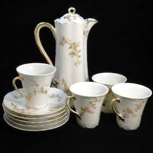 May include: A white porcelain coffee set featuring a tall lidded pot, three cups, and four saucers. Each piece is decorated with delicate pink floral sprays, green leaves, and gold trim on the handles, rims, and the lid finial.