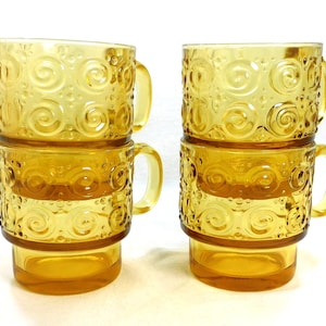 May include: Four amber-colored glass mugs stacked in pairs. Each mug features a textured design of swirls and small dots. The mugs have a handle and a sturdy base, perfect for serving hot beverages.