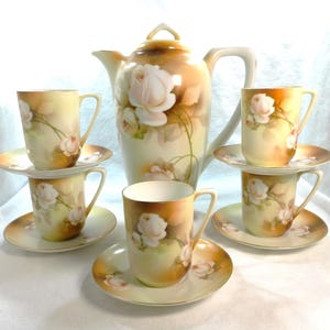 May include: A vintage porcelain tea set with a cream and gold color scheme. The set features a teapot and four cups and saucers. Each piece is decorated with a delicate floral design of white roses.