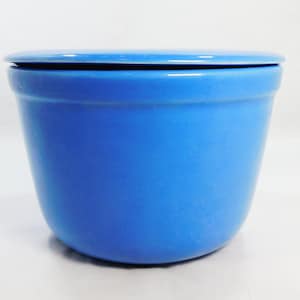 May include: A bright blue ceramic bowl with a matching lid. The bowl has a smooth, rounded exterior with a slightly tapered shape, featuring a wide opening at the top and a flat base. The lid sits flush on top, completing the solid blue design.