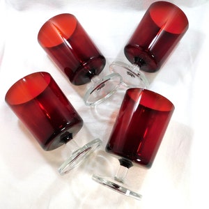 May include: Set of four red glass wine glasses with clear glass stems and bases. The glasses are arranged in a cluster on a white surface.