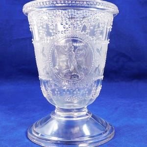 May include: Clear glass goblet with a detailed design. The goblet features a round base, a stem, and a wide bowl. The bowl is decorated with a circular emblem of a figure and floral patterns. The rim is adorned with small, raised dots.