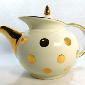 May include: Cream-colored teapot with gold accents and polka dots. The spout, handle, and lid are trimmed in gold. The teapot has a rounded body and a curved spout.
