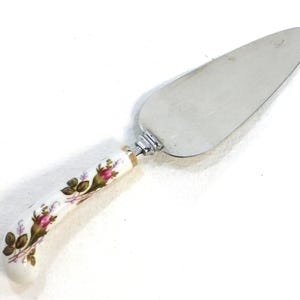 May include: A cake server with a stainless steel blade and a white handle decorated with pink roses and green leaves. The handle is curved and has a silver metal accent where it meets the blade. A classic serving utensil.