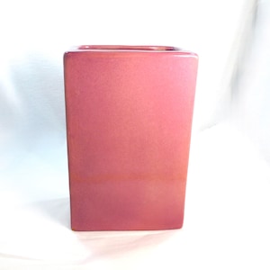 May include: A rectangular ceramic vase with a smooth, glossy finish in a vibrant shade of pink. The vase features clean, straight edges and a wide, open top, showcasing a uniform color throughout the entire exterior surface of the object.