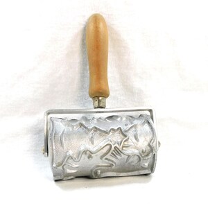 May include: A silver rolling pin with a wooden handle. The rolling pin has a decorative design of a repeating pattern of leaves and flowers.