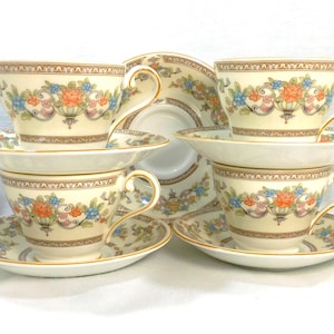 May include: A set of six cream-colored teacups and saucers. Each cup and saucer features a floral design with orange, blue, and yellow flowers. The rims and handles are trimmed with gold. The teacups are stacked in pairs.