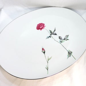 May include: White oval serving platter with a silver rim and a single red rose with green leaves and stems.