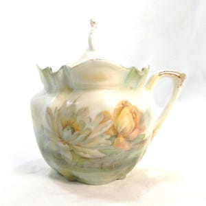 May include: A porcelain sugar bowl with a lid, featuring a scalloped rim and a handle with gold trim. The body is decorated with hand-painted white and yellow water lily flowers against a soft green and cream background with subtle shading.