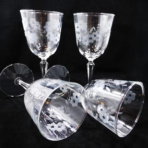 May include: Four clear glass wine glasses featuring an etched floral pattern of small blossoms and delicate vines. Two glasses stand upright on their circular bases, while two are positioned horizontally, showcasing the etched design on the bowls.