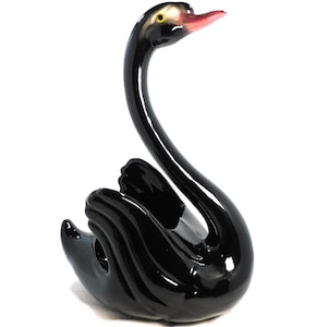 May include: A glossy black swan figurine with a long, curved neck and a gracefully curved body. The swan's beak is a soft pink, and its eye is a small yellow dot. The figurine is a decorative item, likely made of ceramic or porcelain.