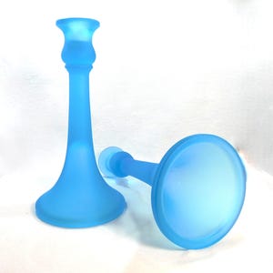 May include: Two frosted blue glass candle holders. One is upright, the other is lying on its side. The candle holders have a classic design with a flared base, a slender stem, and a cup-shaped top to hold a candle.