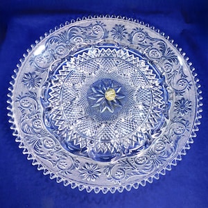 May include: A clear glass serving platter with intricate floral and geometric patterns. The platter has a scalloped edge and a raised center section. A gold-colored sticker is in the center. The design is symmetrical and detailed, suitable for serving appetizers or desserts.