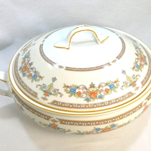 May include: Cream-colored, oval serving dish with a lid. The dish features a floral pattern in blue, orange, and green, accented with gold trim. It has two side handles and a handle on the lid. The dish is likely made of porcelain or ceramic.
