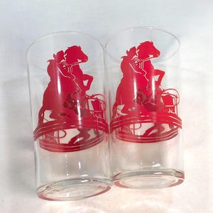 May include: Two clear drinking glasses with a red silhouette design of a horse and rider. The glasses have a cylindrical shape and a red band around the base. The design is repeated on both glasses.