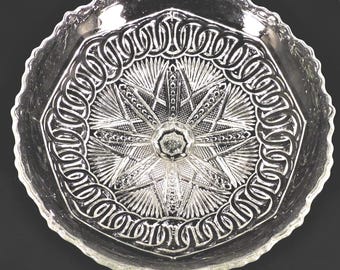 May include: A clear glass serving bowl featuring a central starburst pattern surrounded by a ring of interlocking chain-link motifs. The bowl has a scalloped, decorative edge and a pressed glass design with intricate geometric detailing.