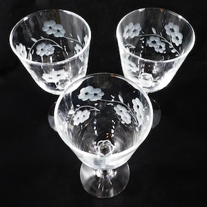 May include: Three clear glass goblets arranged in a triangular formation. Each goblet features a delicate, etched floral pattern with five-petaled flowers and thin, curving stems encircling the upper portion of the bowl and a circular base.