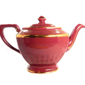 May include: A maroon ceramic teapot with a gold rim and accents. The lid features a textured pattern and a small finial. The handle and spout are curved, and the teapot has a classic design.