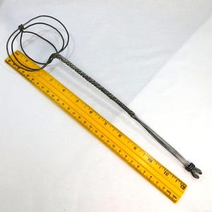 May include: A handmade metal tool with a twisted handle and a looped end. A yellow ruler is placed diagonally across the tool, showing measurements in inches. The tool appears to be made of dark metal.