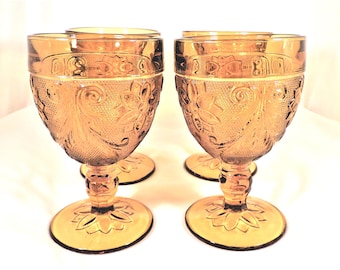 May include: Set of four amber-colored goblets. Each goblet features a floral and swirl design on the bowl and a detailed stem. The base of each goblet has a flower design. The goblets are made of glass.
