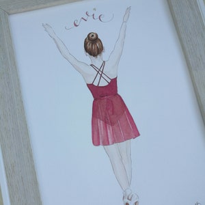 May include: A watercolor painting of a ballerina in a red dress with her arms raised above her head. The painting is framed in a light wood frame. The name 'Evie' is written in cursive at the top of the painting.