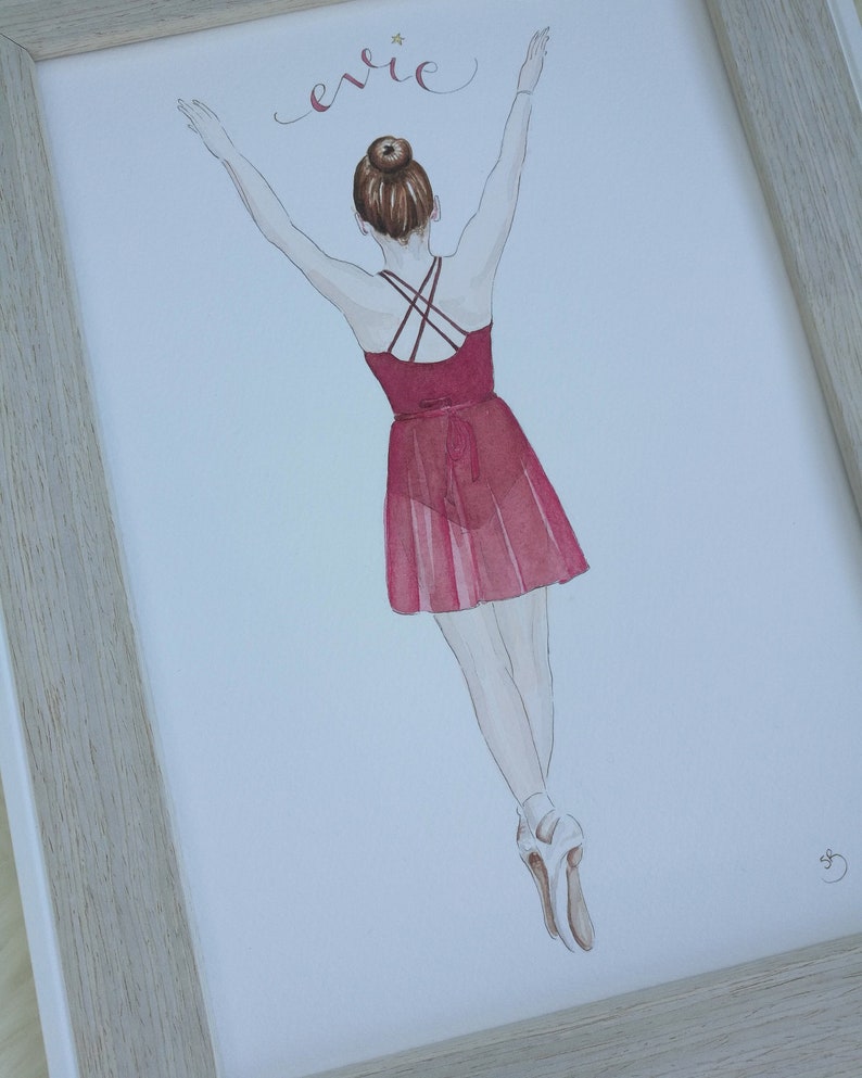 May include: A watercolor painting of a ballerina in a red dress with her arms raised above her head. The painting is framed in a light wood frame. The name 'Evie' is written in cursive at the top of the painting.