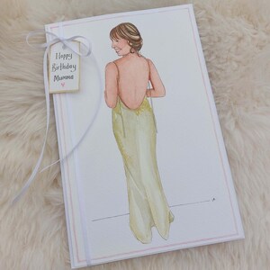 May include: A white card with a pink border. The card has a watercolor illustration of a woman in a light green dress. The card has a tag with the text 'Happy Birthday Mumma'.