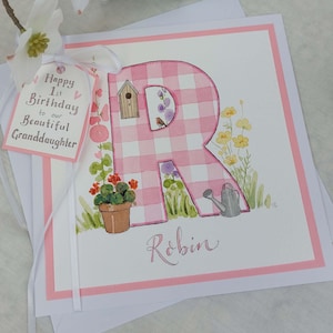 Peut inclure: Une lettre R en vichy rose et blanc avec une maison d'oiseau, un rouge-gorge, un arrosoir et des fleurs. La carte dit "Happy 1st Birthday to our beautiful Granddaughter" et "Robin".