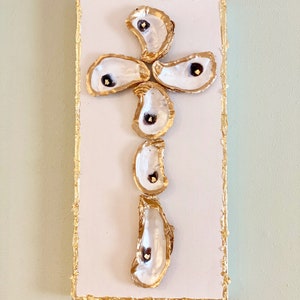 May include: A white canvas with a gold-leaf border features a cross design made from oyster shells. The shells are white with gold edges and a dark spot in the center. The artwork is a unique, coastal-themed piece.