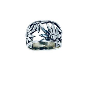 May include: A silver-colored ring with a floral daisy design. The ring features cut-out daisy shapes around the band. The ring is shown against a white background. The ring is a wide band style.